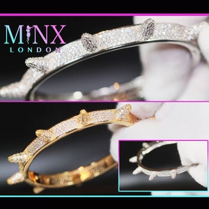 Może przedstawiać: Srebrna i złota bransoletka z kolczastym wzorem, inkrustowana małymi, błyszczącymi kamieniami. Obraz przedstawia dwie wersje bransoletki, z nazwą marki "MINX LONDON" wyświetlaną u góry.