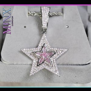 Könnte beinhalten: Eine silberfarbene Stern-Anhänger-Halskette mit einem kleineren rosa Stern in der Mitte. Der äußere Stern ist mit klaren Kristallen besetzt. Der Anhänger hängt an einer silbernen Kette. Der Text "MINX LONDON" ist sichtbar.