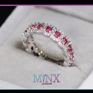 May include: A silver ring with a continuous row of pink gemstones and smaller clear stones. The ring is set in a silver band and is displayed in a gray jewelry box. The brand name "MINX LONDON" is at the bottom.