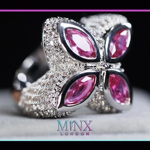 Op de afbeelding: Een zilveren ring met een bloemmotief met vier roze edelstenen. De ring is bezet met kleine, heldere stenen en de merknaam "MINX LONDON" is onderaan zichtbaar.