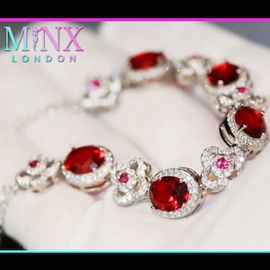 Puede incluir: Un brazalete de plata con piedras preciosas ovaladas rojas rodeadas de piedras preciosas más pequeñas y transparentes. El brazalete está sobre una superficie blanca.