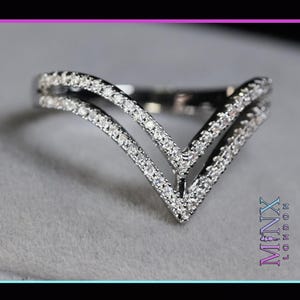 Puede incluir: Un anillo plateado con un diseño en forma de V. El anillo presenta dos bandas paralelas, cada una incrustada con pequeñas piedras brillantes. El anillo se muestra sobre un fondo neutro, resaltando su estilo elegante y moderno.