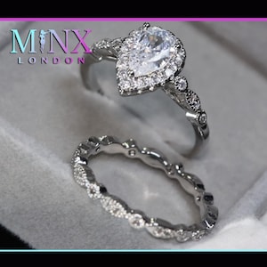 May include: Two silver rings. The top ring features a pear-shaped, clear gemstone surrounded by smaller stones. The band is adorned with additional small stones. The second ring is a band with a repeating pattern of small, clear stones. The rings are displayed on a light gray surface.