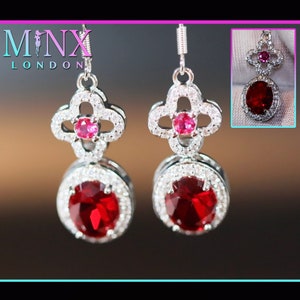 May include: Silver-toned drop earrings featuring a clover-shaped top with a small pink gemstone center and a larger, oval-shaped red gemstone at the bottom, surrounded by small white stones. The earrings have a hook closure. The brand name "MINX LONDON" is visible.
