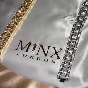 May include: Two rhinestone bracelets, one gold-toned and one silver-toned, are displayed on a white satin bag. The bag features the brand name "MONX LONDON". The bracelets are rectangular with multiple sparkling stones.