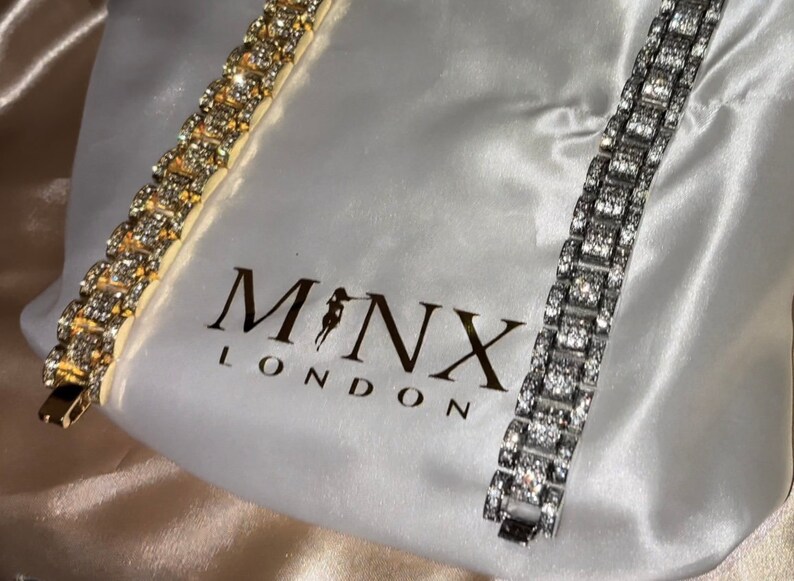 May include: Two rhinestone bracelets, one gold-toned and one silver-toned, are displayed on a white satin bag. The bag features the brand name "MONX LONDON". The bracelets are rectangular with multiple sparkling stones.