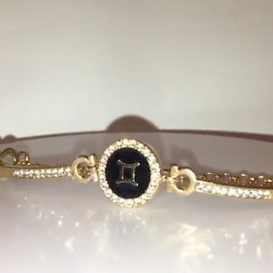 May include: Gold bracelet with a black enamel charm featuring the Gemini zodiac symbol. The charm is surrounded by clear rhinestones.
