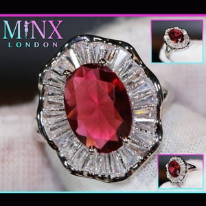 May include: A silver ring with a large, oval-shaped, red gemstone surrounded by a halo of smaller, clear gemstones. The ring is set in a silver band.