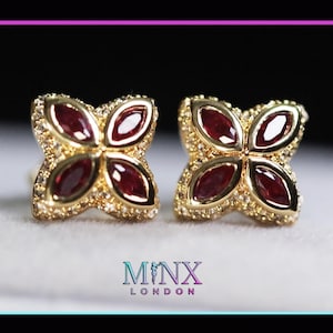 May include: A pair of gold-colored stud earrings. Each earring features a four-petal design with a central, oval-shaped, deep red gemstone in each petal. The petals are surrounded by small, clear gemstones. The earrings are set against a soft, neutral background.