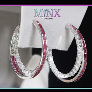 Peut inclure: Boucles d'oreilles créoles argentées avec des rangées alternées de pierres précieuses claires et rouges. Les boucles d'oreilles ont un design superposé. Le nom de la marque "MINX LONDON" est affiché en haut.