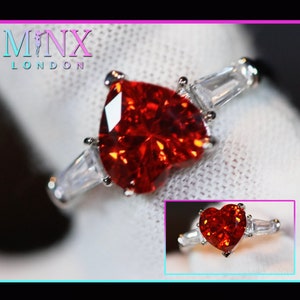 Peut inclure: Une bague en argent ornée d'une pierre précieuse rouge vif en forme de cœur. La bague est flanquée de deux pierres précieuses rectangulaires et transparentes. Le texte "MNX LONDON" est visible en haut à gauche.