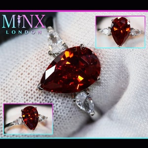 May include: A pear-shaped red gemstone ring with smaller pear-shaped clear gemstones on either side. The ring is set in silver.