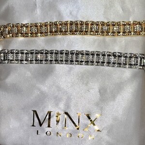 May include: Two rhinestone bracelets, one gold and one silver, rest on a white satin bag. The bracelets feature a repeating pattern of rectangular stones. The bag has the gold text "MINX LONDON".