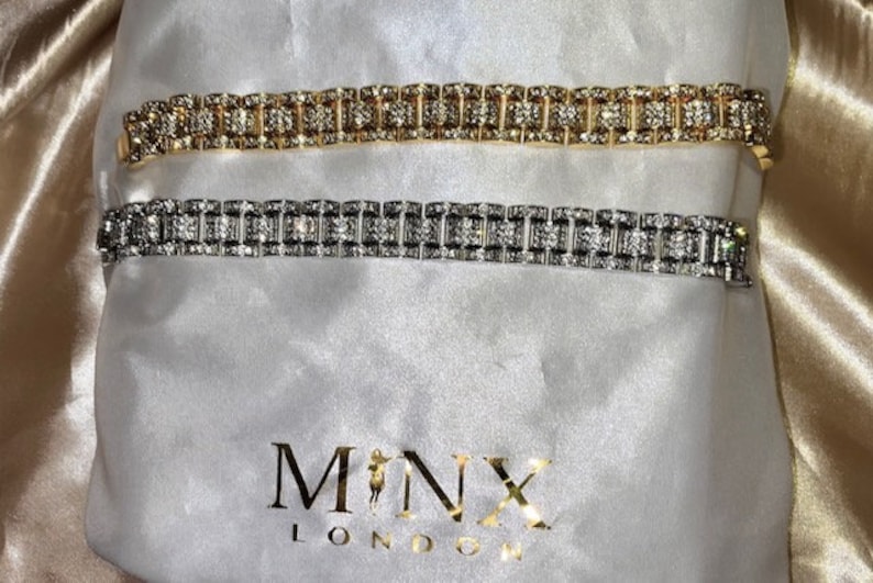 May include: Two rhinestone bracelets, one gold and one silver, rest on a white satin bag. The bracelets feature a repeating pattern of rectangular stones. The bag has the gold text "MINX LONDON".