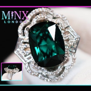 May include: A large emerald green gemstone ring with a silver setting. The ring is surrounded by smaller diamonds. The ring is on a white surface. MINX LONDON is written in the top left corner of the image.
