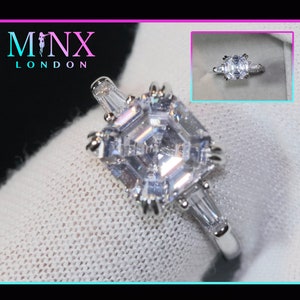 May include: A silver ring featuring a large, clear, square-shaped diamond. The diamond has a step-cut design and is surrounded by smaller diamonds. The ring is displayed on a white fabric background.