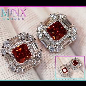 May include: A pair of silver earrings with a red square gemstone surrounded by smaller white gemstones. The earrings have a square shape with a decorative border of smaller white gemstones.