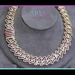 Puede incluir: Un collar de cadena cubana dorado con piedras preciosas rojas y blancas. El collar se exhibe sobre un busto gris. El cierre es rectangular y presenta piedras preciosas rojas. La marca "MNX LONDON" está en la parte superior central.