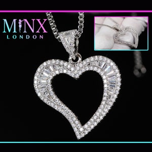 Puede incluir: Un collar de plata con un gran colgante de corazón abierto incrustado con piedras de circonita cúbica transparentes. El colgante tiene una doble fila de piedras alrededor del borde y una sola fila de piedras alrededor de la forma del corazón. El collar está en una cadena de plata.