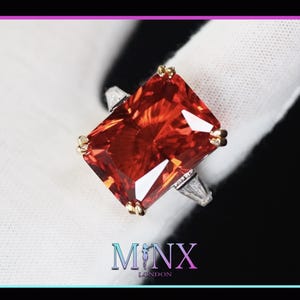 May include: A large, rectangular, red gemstone ring set in a silver band with small, clear gemstones on the sides. The ring is on a white surface.