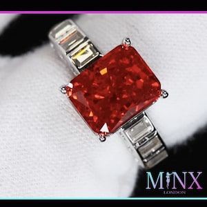 May include: A silver ring with a large, red, square-shaped gemstone surrounded by smaller clear gemstones. The ring is on a white surface. MINX LONDON is printed on the bottom right corner of the image.