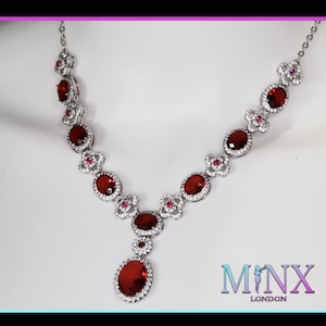 Puede incluir: Un collar de plata con un gran colgante de piedra roja y piedras rojas más pequeñas a lo largo de la cadena. El collar está adornado con piedras transparentes más pequeñas. El nombre de la marca "MINX LONDON" es visible en la esquina inferior derecha.