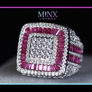 Könnte beinhalten: Ein quadratischer, silberfarbener Ring mit einem zentralen Quadrat aus kleinen, klaren Steinen. Der Ring ist von einer Reihe rosa Steine umrahmt, und das Band ist mit klaren Steinen besetzt. Der Markenname "MINX LONDON" ist zu sehen.
