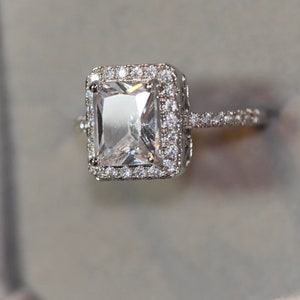 May include: A silver ring featuring a large, clear, square-shaped gemstone surrounded by a halo of smaller diamonds. The ring has a delicate band with smaller diamonds.