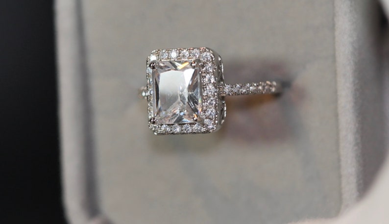 May include: A silver ring featuring a large, clear, square-shaped gemstone surrounded by a halo of smaller diamonds. The ring has a delicate band with smaller diamonds.