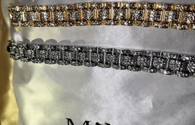 May include: Two rhinestone-studded bracelets. One is gold-toned, the other silver-toned. The bracelets have a watch band style with rectangular links. The background is a soft, white fabric.