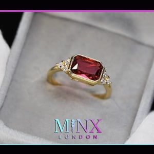 May include: A gold ring with a large, rectangular, red gemstone surrounded by smaller, clear gemstones. The ring is in a small, gray box with the text "MINX LONDON" on the bottom.