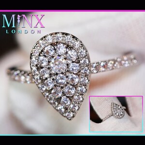 Puede incluir: Primer plano de un anillo plateado con un grupo de pequeñas piedras blancas brillantes en forma de pera. La banda del anillo también está adornada con pequeñas piedras transparentes. La marca "MINX LONDON" es visible.