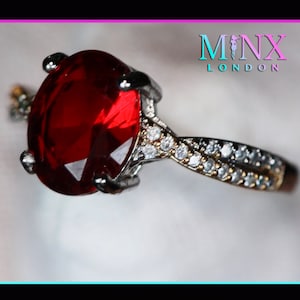 May include: A silver ring featuring a large red oval gemstone and a band of smaller white gemstones. The ring is displayed against a white backdrop. The words MINX LONDON are visible in the top right corner of the image.