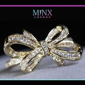 May include: A gold-colored bow ring encrusted with numerous small, clear gemstones. The ring's design features a detailed bow shape, with the gemstones set along the curves and center. The brand name "MINX LONDON" is displayed at the top.