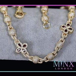 Puede incluir: Collar dorado con diseño floral y gemas rojas. El collar está incrustado con pequeñas piedras brillantes y tiene un cierre. El diseño incluye eslabones interconectados y detalles en forma de flor. La marca es 'MINX LONDON'.