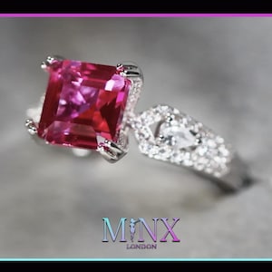 May include: A silver ring featuring a large, square-shaped, pink gemstone surrounded by smaller, clear gemstones. The ring is set in a delicate, intricate design. The brand name "MINX LONDON" is visible below the ring.