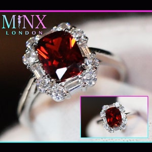 Puede incluir: Un anillo de plata con una gran piedra preciosa roja cuadrada rodeada de piedras preciosas más pequeñas y transparentes. El anillo está engastado en un diseño delicado y ornamentado. El nombre de la marca "MINX LONDON" es visible en el fondo.