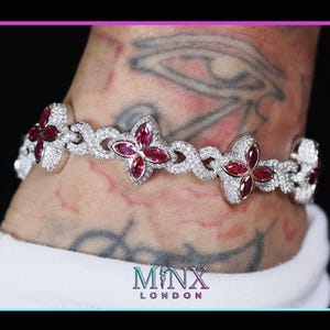 Może przedstawiać: Srebrna bransoletka z kwiatowym wzorem. Każdy kwiat ma czerwony kamień szlachetny w środku, otoczony przezroczystymi kryształami. Bransoletka jest na nadgarstku z tatuażami. Widoczna jest nazwa marki "MINX LONDON".