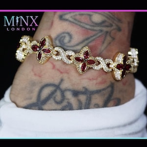 Può includere: Bracciale dorato con motivi floreali a quattro petali con pietre rosse al centro e cristalli trasparenti. Il bracciale ha un design intricato e intrecciato. Il marchio "MINX LONDON" è visibile.