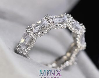 May include: A silver ring with a continuous design of rectangular and round-cut clear stones. The ring is set on a white surface. The brand name "MINX LONDON" is displayed in a teal and purple font at the bottom of the image.