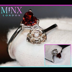 May include: A silver ring featuring a red heart-shaped stone and a white rose design. The ring is displayed on a white background.