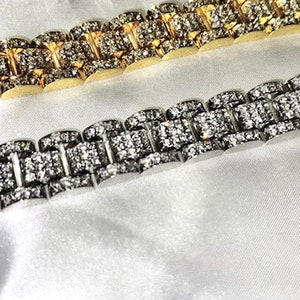 May include: Two luxury bracelets, one gold-toned and one silver-toned, both encrusted with sparkling gemstones. The bracelets are displayed on a white, silky surface, showcasing their intricate design and high-end appeal.
