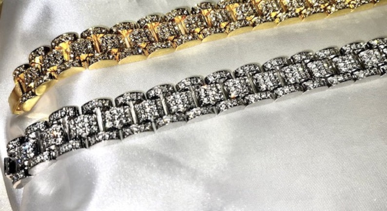 May include: Two luxury bracelets, one gold-toned and one silver-toned, both encrusted with sparkling gemstones. The bracelets are displayed on a white, silky surface, showcasing their intricate design and high-end appeal.