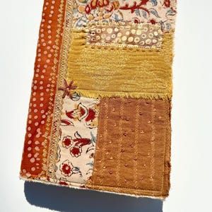 May include: A handmade journal with a patchwork design. The cover features various fabrics in shades of orange, yellow, and cream, with floral and dotted patterns. A small, star-shaped embellishment adds detail to the spine.