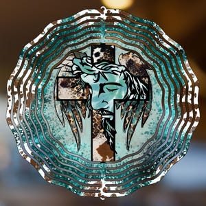 May include: A layered metal wind spinner featuring a religious design. The central image depicts a cross with a face and wings, set against a turquoise and brown background. The outer layers have a wavy pattern in shades of brown, white, and teal.