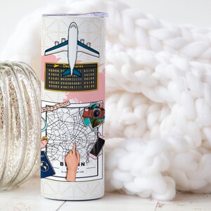 May include: A white travel-themed tumbler with a pink and gold design. The tumbler features a plane, a map of Berlin, a camera, and a passport. The text "Berlin" is visible on the tumbler.