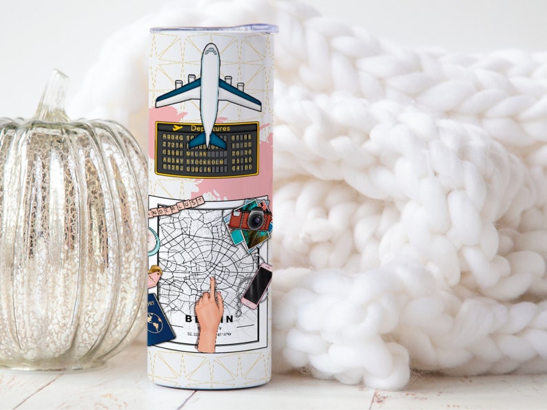 May include: A white travel-themed tumbler with a pink and gold design. The tumbler features a plane, a map of Berlin, a camera, and a passport. The text "Berlin" is visible on the tumbler.