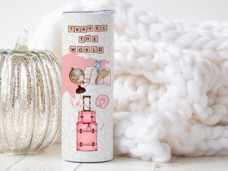 May include: A pink and white travel tumbler with the words "TRAVEL THE WORLD" in black letters. The tumbler features a globe, a map, a suitcase, and a neck pillow.