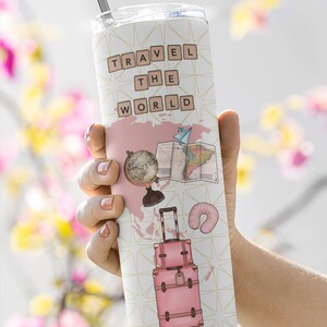 May include: A pink travel tumbler with a white background and gold geometric pattern. The tumbler features the words "TRAVEL THE WORLD" in brown letters, a globe, a map, a suitcase, and a neck pillow.