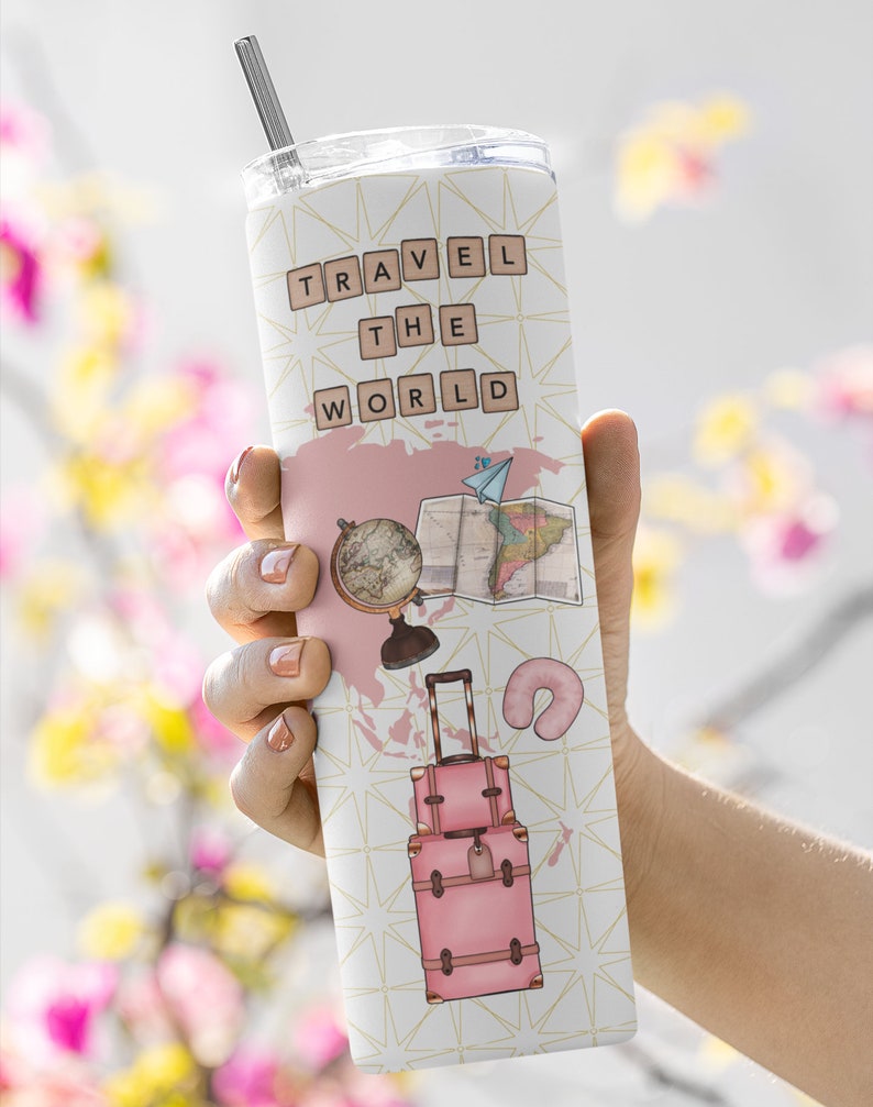 May include: A pink travel tumbler with a white background and gold geometric pattern. The tumbler features the words "TRAVEL THE WORLD" in brown letters, a globe, a map, a suitcase, and a neck pillow.